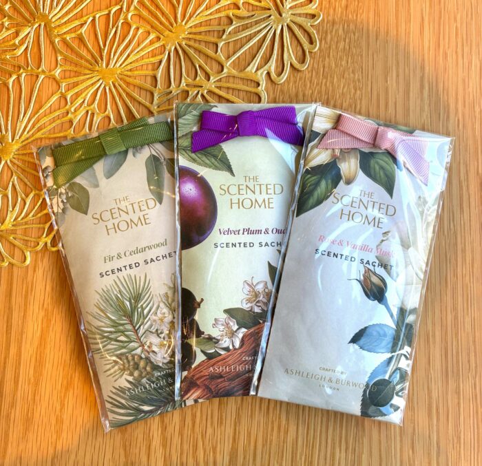 Assorted scented sachets with botanical illustrations and purple, green, and pink ribbons on a wooden background.